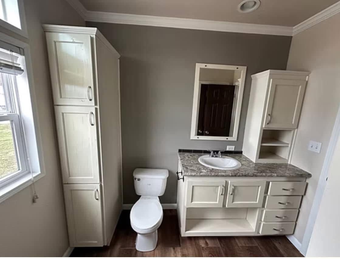 Modern bathroom with a white toilet beside a beige vanity featuring a marble countertop and sink. There are cabinet units, a mirror, and a window with blinds, all in a neutral color scheme. The wood floor adds warmth to the clean, organized space.