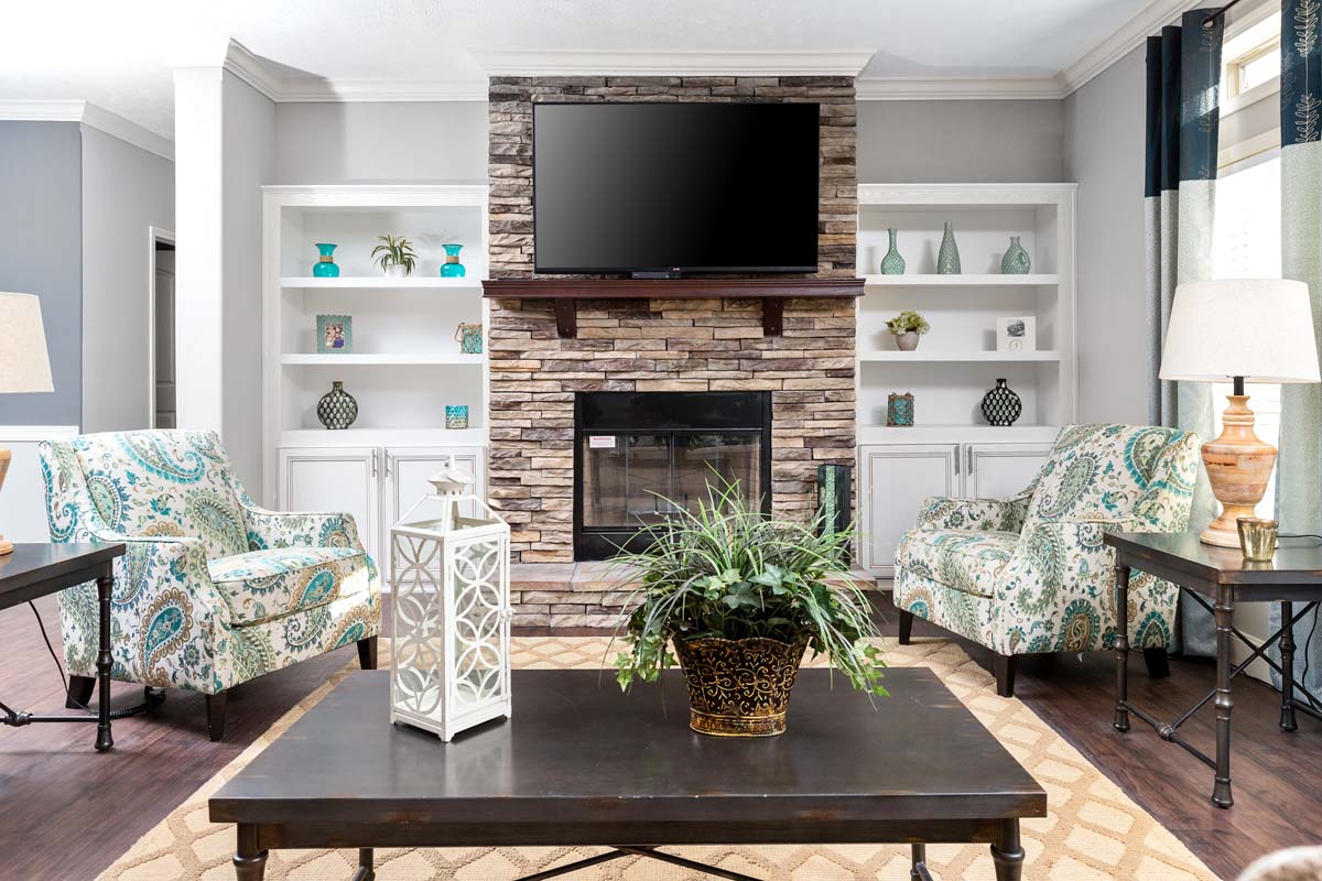 Living room with stone fireplace, flanked by white shelves. Two patterned armchairs face a dark coffee table with a plant. A TV hangs above the mantle.