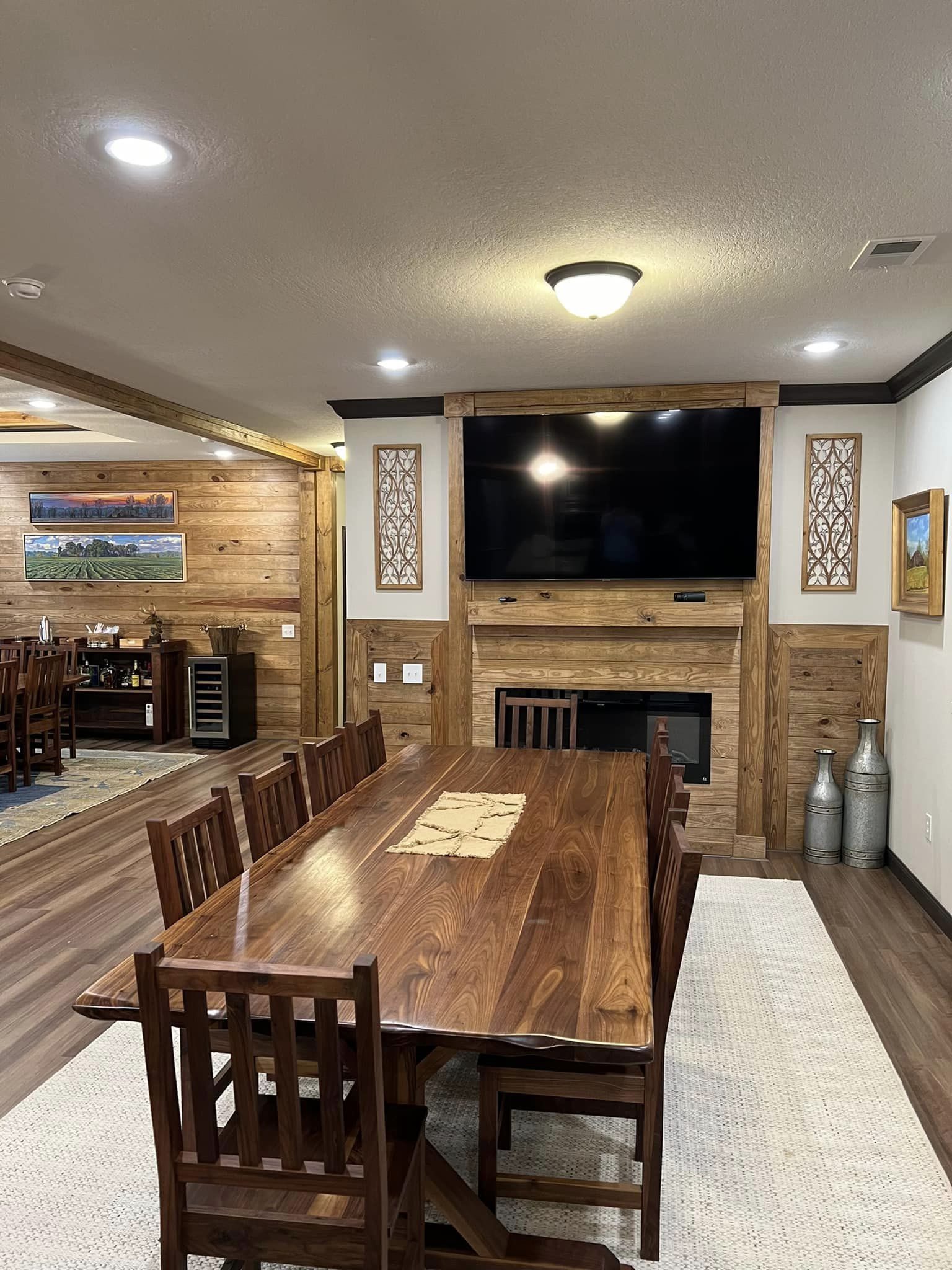 Wooden dining room with rustic decor featuring a long table, chairs, large TV, and fireplace. Warm tones create a cozy and inviting atmosphere.