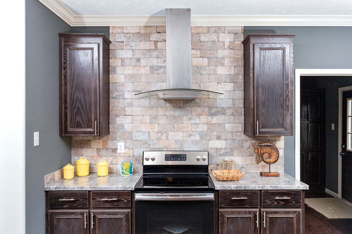Modern kitchen featuring dark wood cabinets, a stainless steel stove with a vent hood, rustic brick backsplash, and yellow decorative jars. Cozy and contemporary.