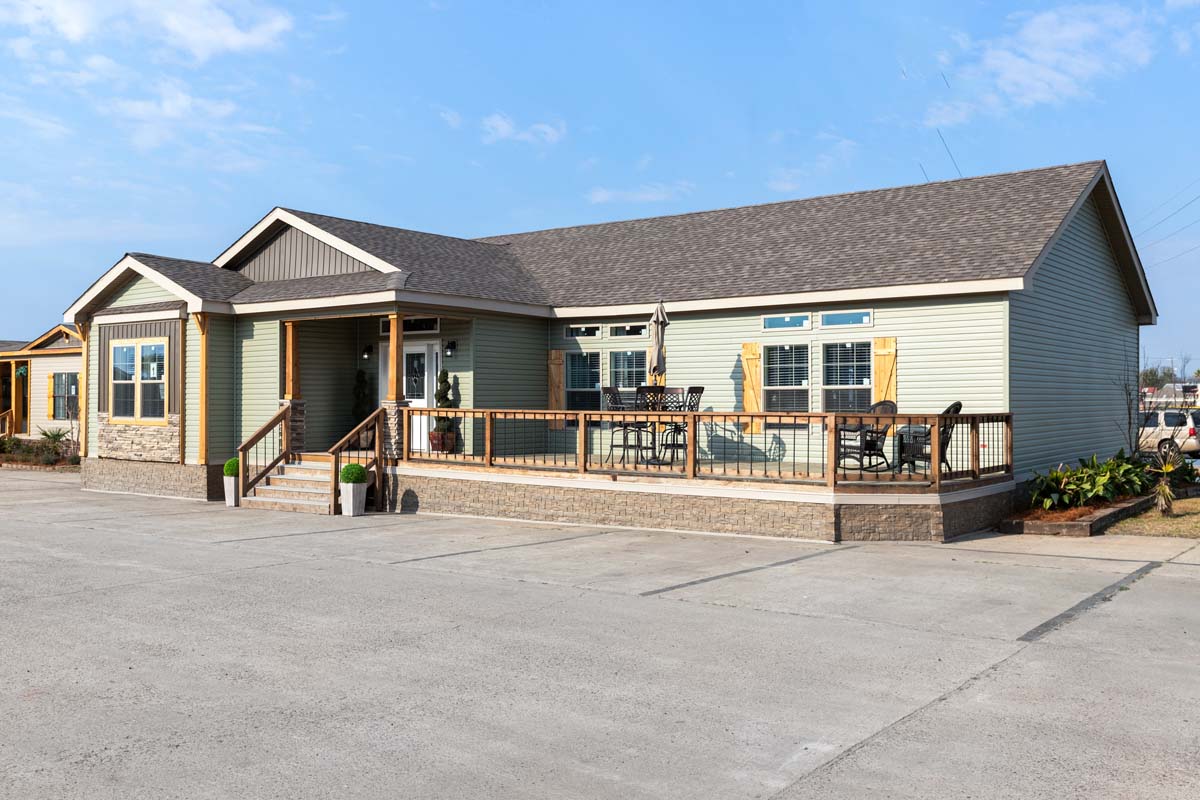 Single-story house with light green siding and stone accents. It features a spacious wooden deck with patio furniture, set on a sunny day.