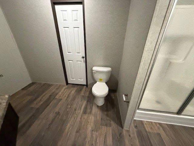 Bathroom with wood-look flooring featuring a toilet next to a narrow door. A shower stall is partially visible, creating a neat, minimalistic ambiance.