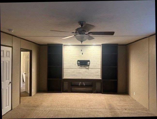 An empty room with beige walls and carpet, featuring a ceiling fan. A TV mount on a white brick wall is flanked by dark, built-in shelves.