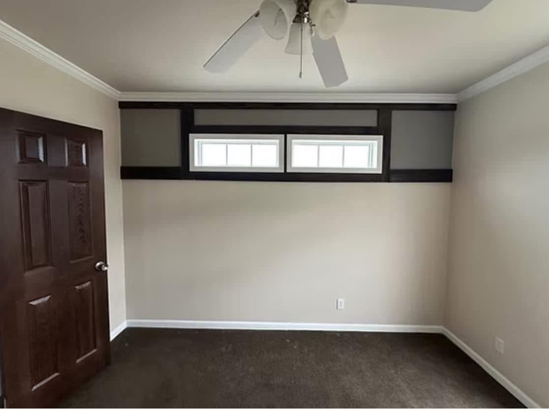 Empty room with beige walls, dark brown carpet, a wooden door on the left, small rectangular windows high on the wall, and a ceiling fan.