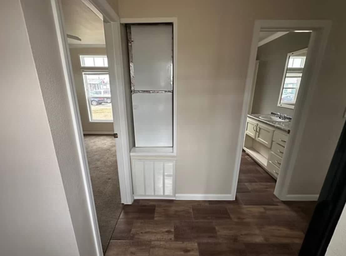 Narrow hallway with two doorways, one leading to a carpeted room, the other to a bathroom with wood flooring and a sink. Bright, neutral tones.