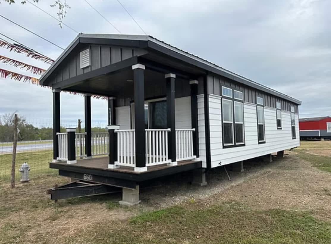 Small white and black prefab home with a front porch, supported by black posts, sits on grass near a road. Overcast sky and calm surroundings.
