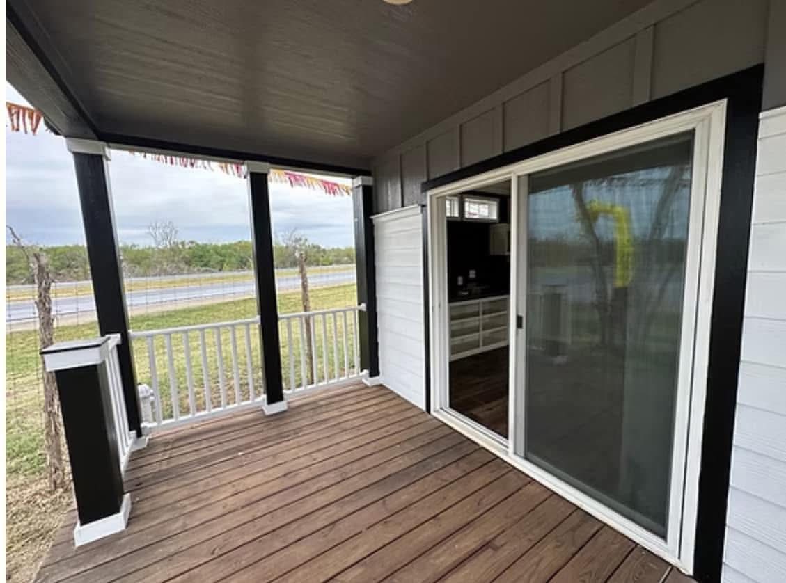 Covered wooden porch with sliding glass door, white railings, and dark trim. Overlooks a grassy yard and road under a cloudy sky. Calm ambiance.