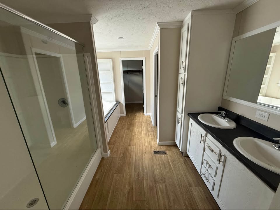 A spacious bathroom with wood flooring, double sinks on a black countertop, a large wall mirror, a glass shower, and an open walk-in closet.