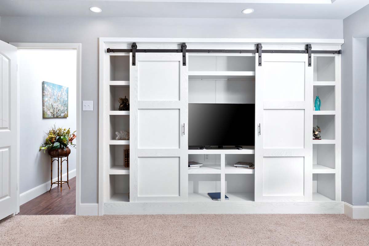 Built-in shelving unit with white sliding barn doors flanking a central TV. Open shelves display books and decor. Soft, carpeted room with bright light.