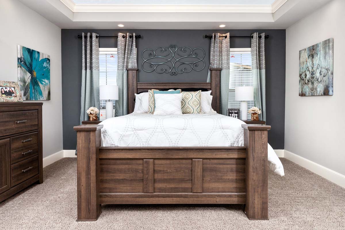 A serene bedroom with a wooden bedframe, white bedding, and decorative pillows. Two side tables with lamps flank the bed, set against gray walls and draped windows.
