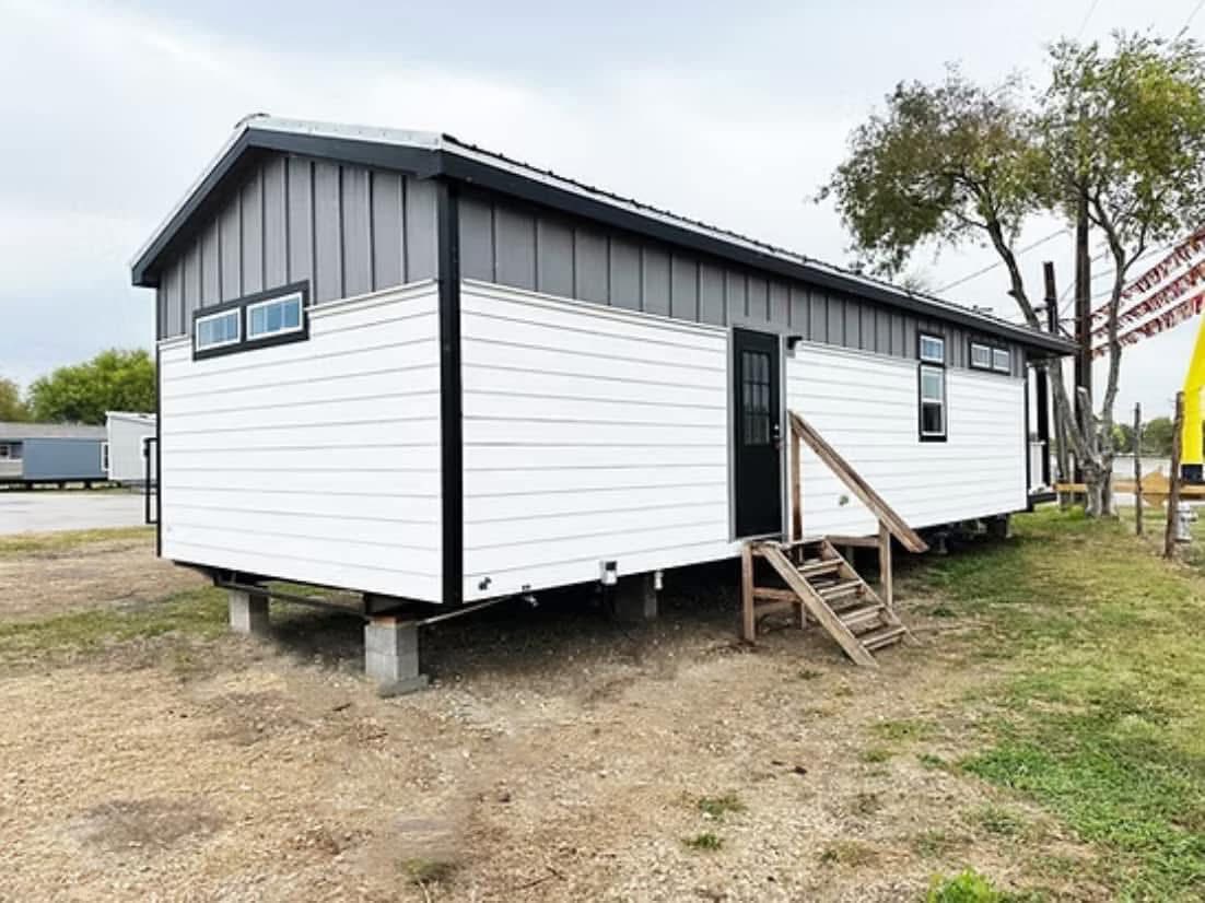 A modern, rectangular tiny house with white siding and black accents sits elevated on blocks. Wooden steps lead to a black door, set against a grassy backdrop.