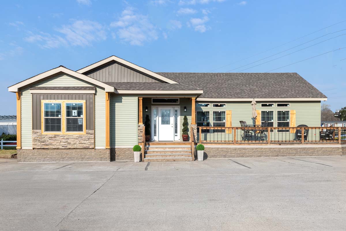 A modern, single-story house with light green siding and stone accents, featuring a welcoming front porch with potted plants, under a clear blue sky.
