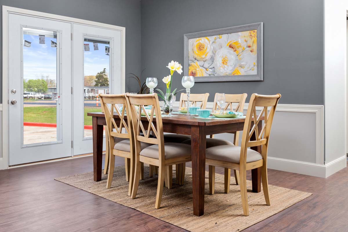 A dining room features a wooden table set with floral decor and turquoise dishes, surrounded by six chairs on a textured rug. A floral painting hangs on a gray wall; light streams in through glass doors, creating a cozy, inviting ambiance.