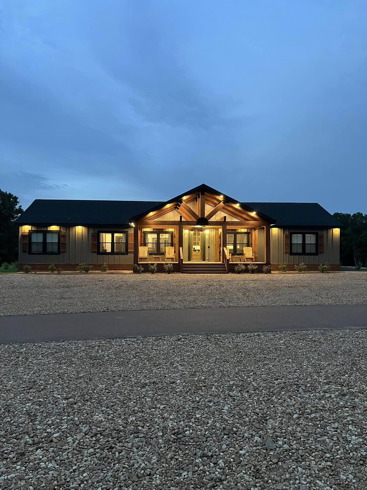 A warmly lit, single-story house with wooden accents and large windows sits on a gravel driveway at dusk, exuding a welcoming and cozy vibe.