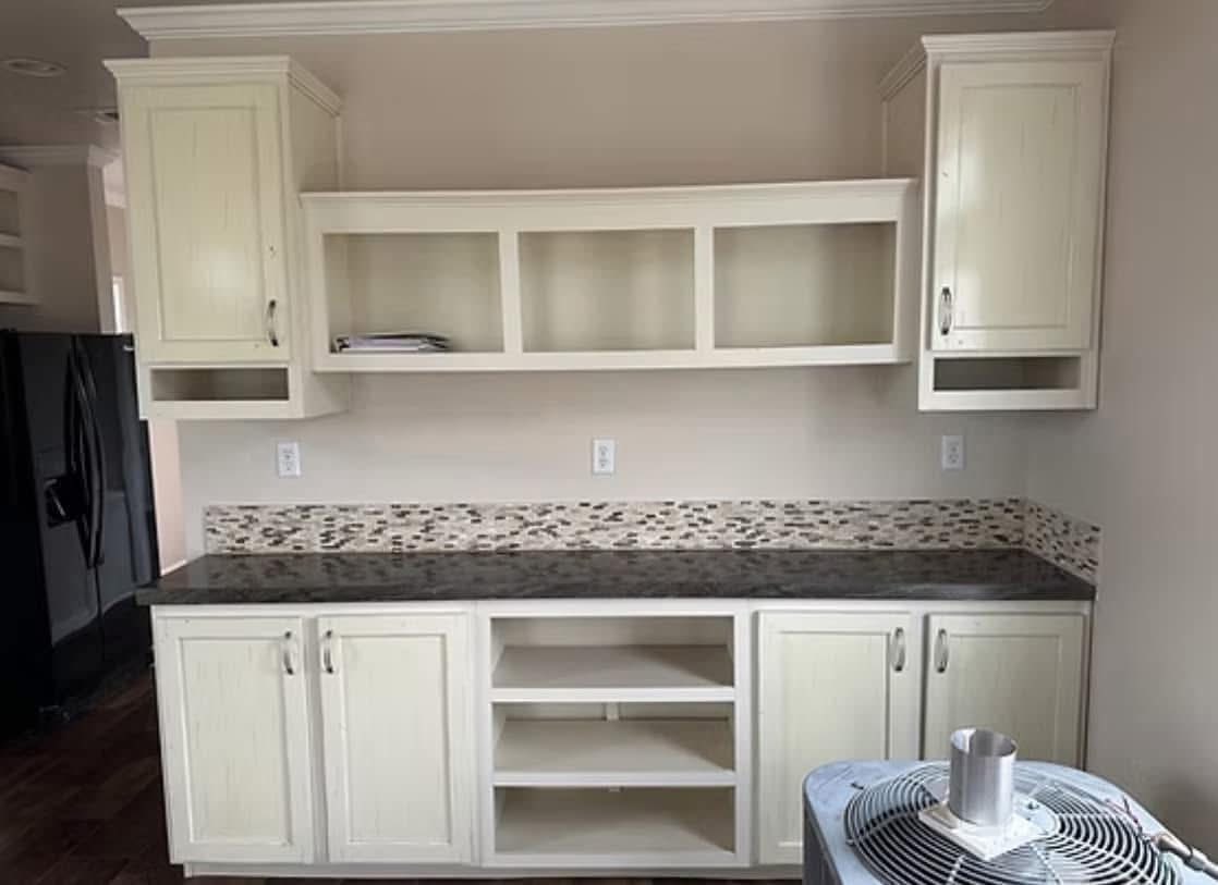 Cream-colored kitchen cabinets with a dark countertop. Mosaic tile backsplash adds contrast. Open shelves hold a magazine, creating a tidy look.