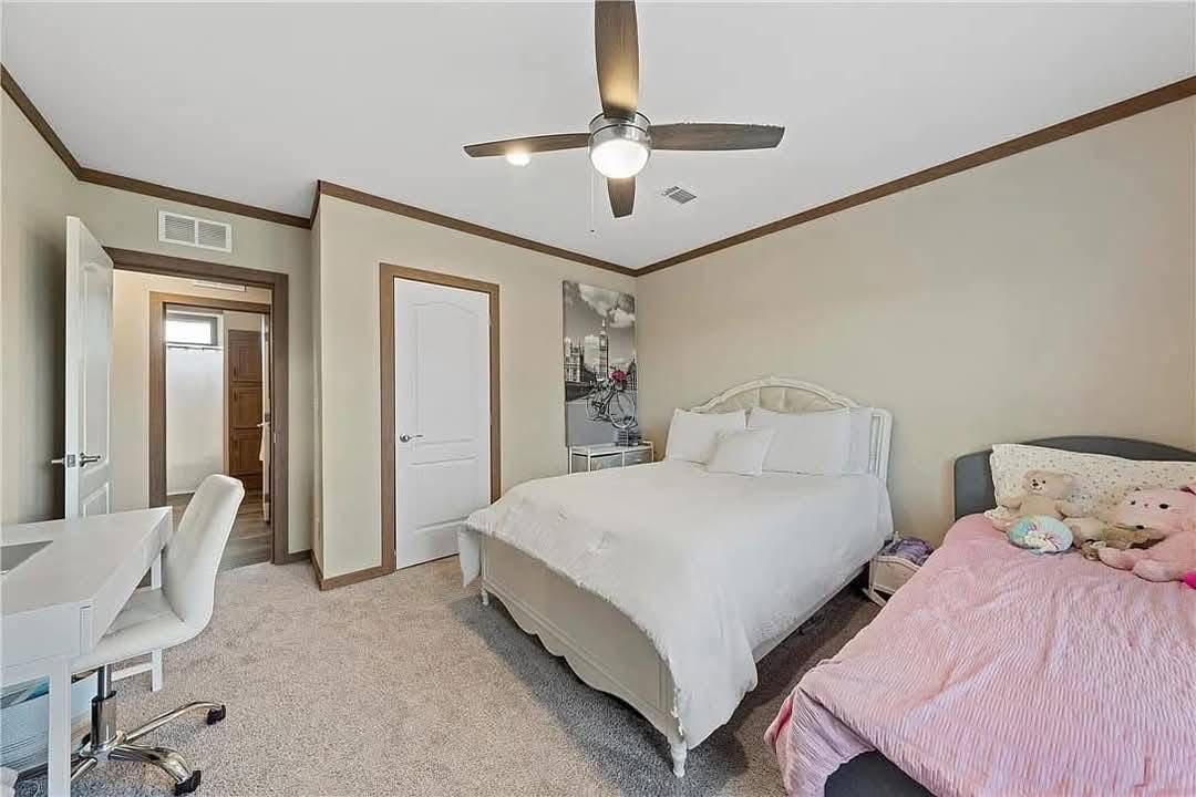 Cozy bedroom with white bed and pink bedding, toys on the side. A modern ceiling fan, desk, and wall art. Neutral tones create a calm, inviting atmosphere.
