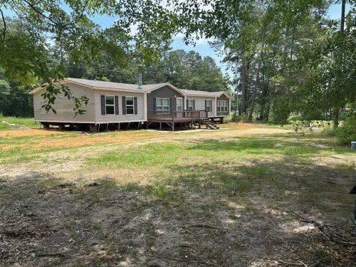 A beige double-wide mobile home sits in a grassy, wooded area under a clear sky, conveying a peaceful, rural atmosphere.