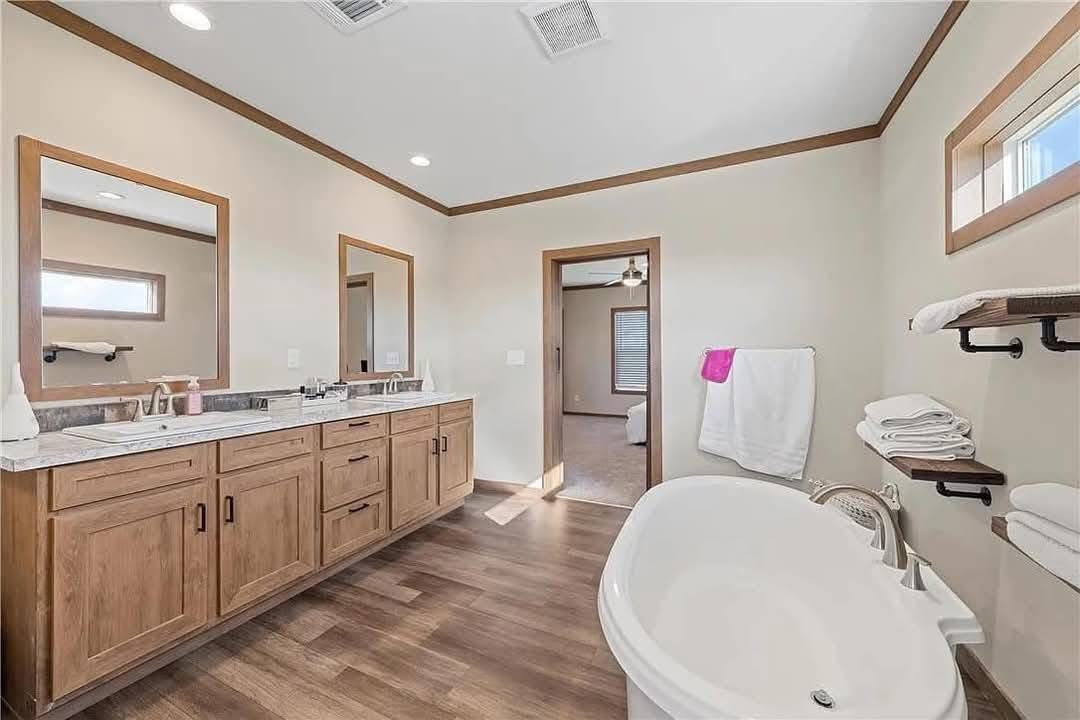 Spacious bathroom with a wooden double vanity, two mirrors, and a white bathtub. Towels hang neatly, and a doorway leads to a bedroom, exuding a calm, modern vibe.