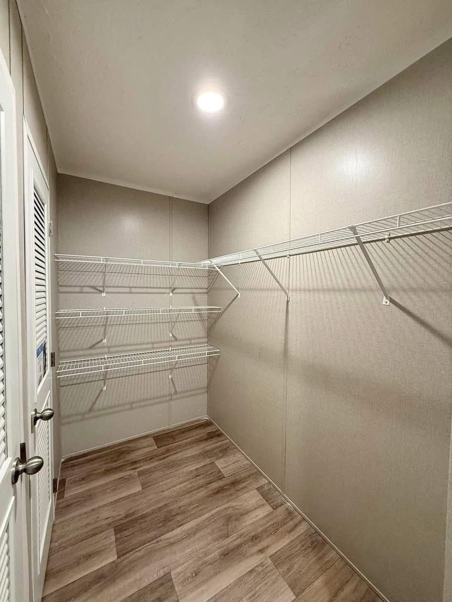 Minimalist walk-in closet with beige walls, a wooden floor, and white wire shelving on two walls under a bright ceiling light. Clean, organized feel.