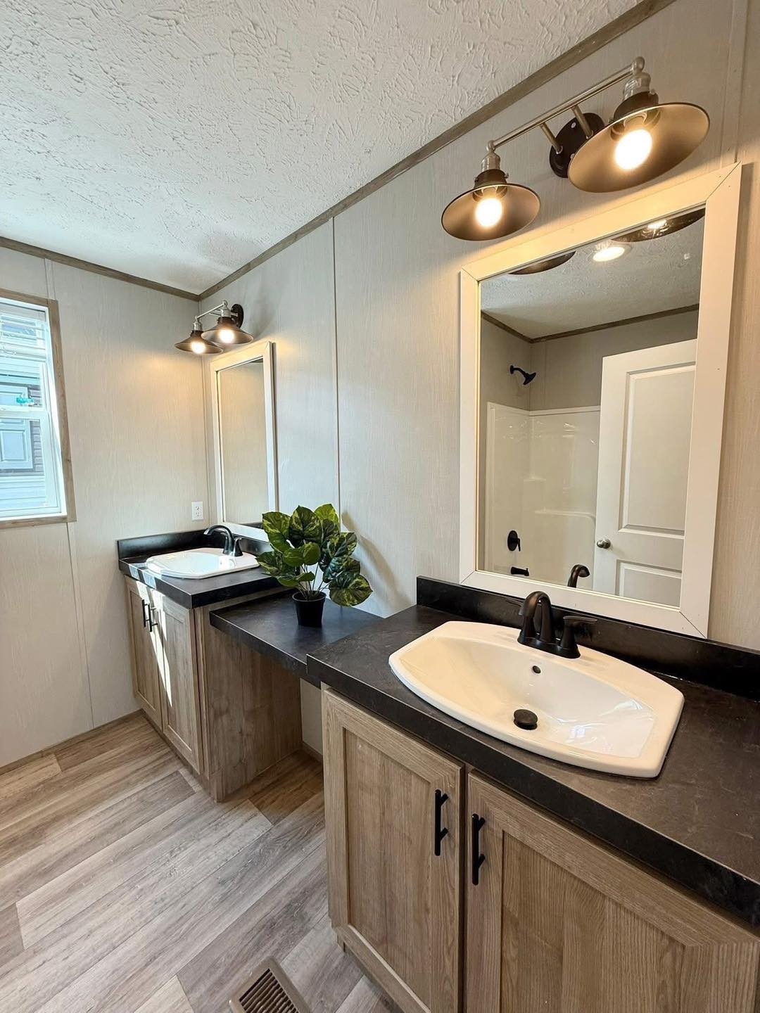 Modern bathroom with two wood vanities and black counters, each with a sink and mirror. A potted plant decorates the space, under warm overhead lights.