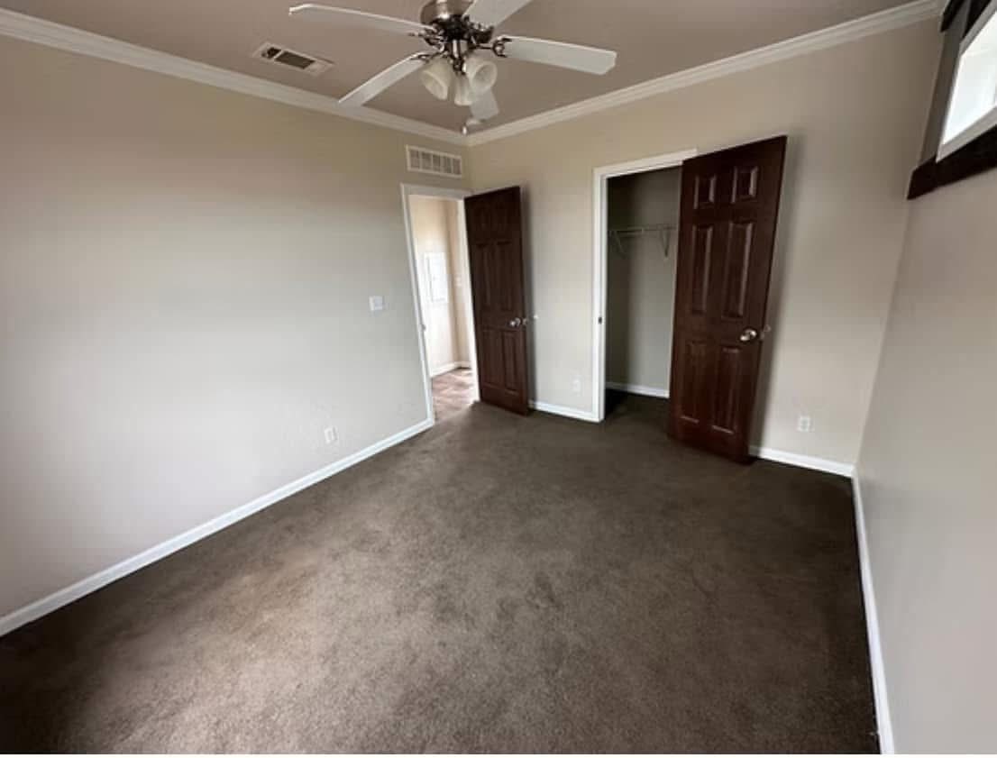 An empty room with beige walls and brown carpet. A ceiling fan hangs above. Two dark wooden doors reveal a closet and hallway, creating a neutral, calm atmosphere.