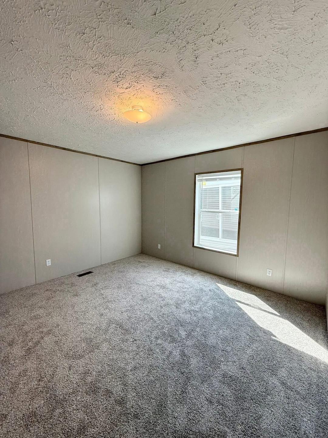 A small, empty room with beige walls and a white ceiling featuring a single ceiling light. A window on the right lets in sunlight, casting shadows on the gray carpeted floor. The room feels simple and clean.