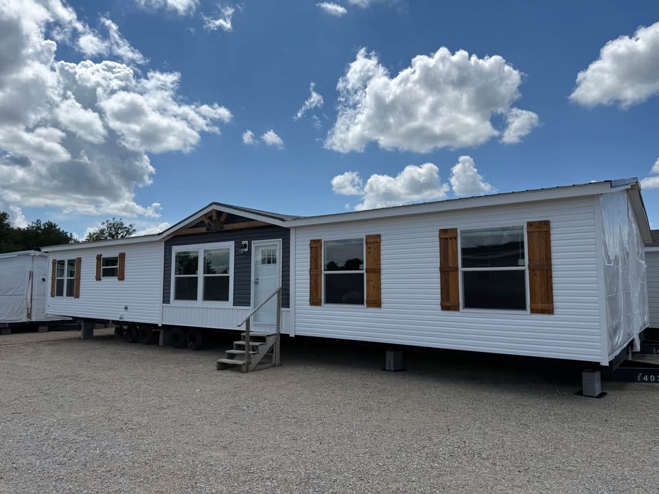 A large white mobile home with wooden shutters sits on a gravel lot under a blue sky with fluffy clouds. The home has a gabled roof and front steps.