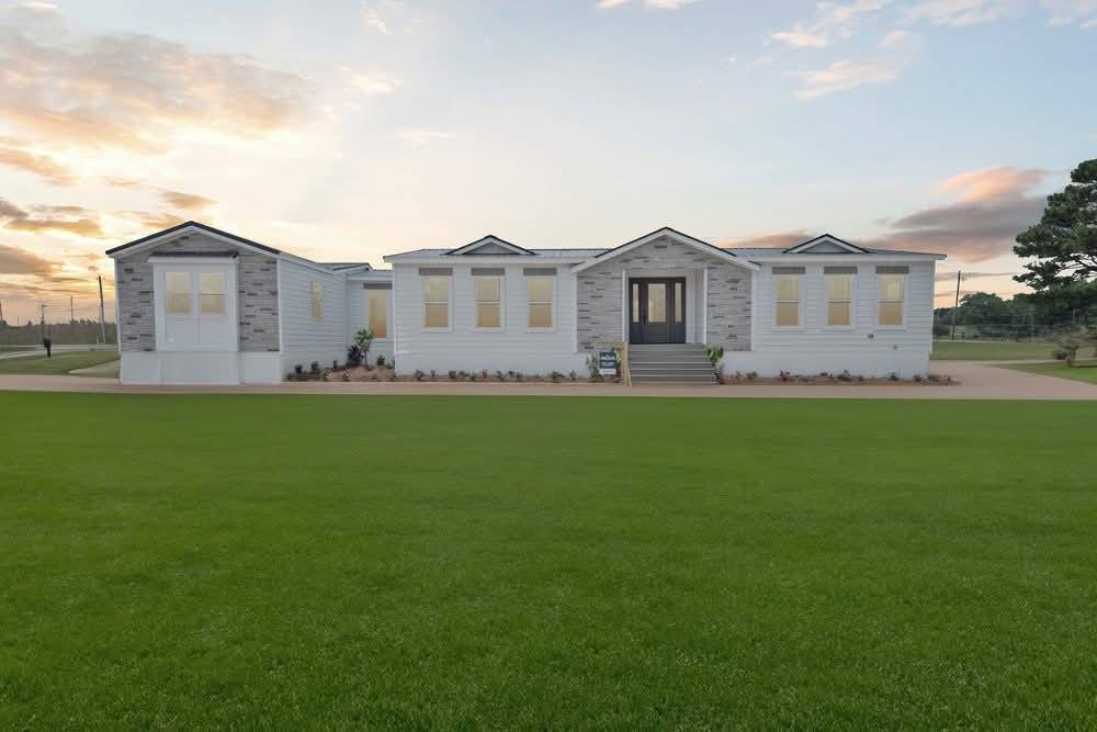 A modern, single-story white house with a central doorway and large windows sits on a vast, lush green lawn under a sunset sky with soft clouds.