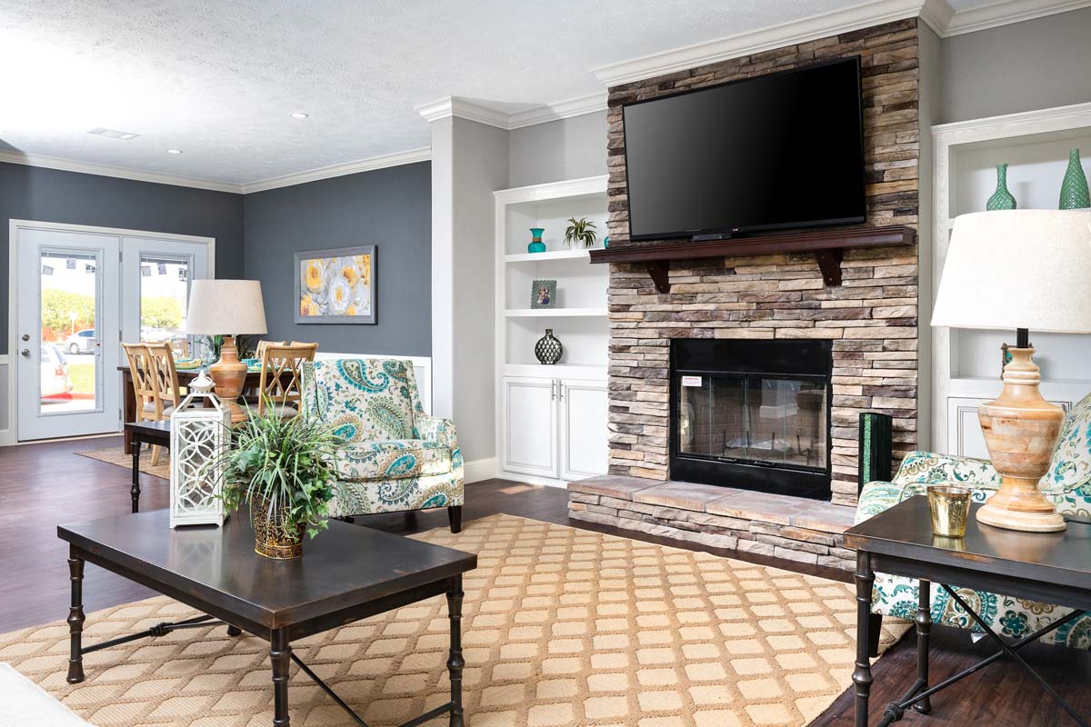 Stylish living room with a stone fireplace, wall-mounted TV, patterned chairs, and dark wood tables. Bright cushions contrast with gray walls, exuding warmth.
