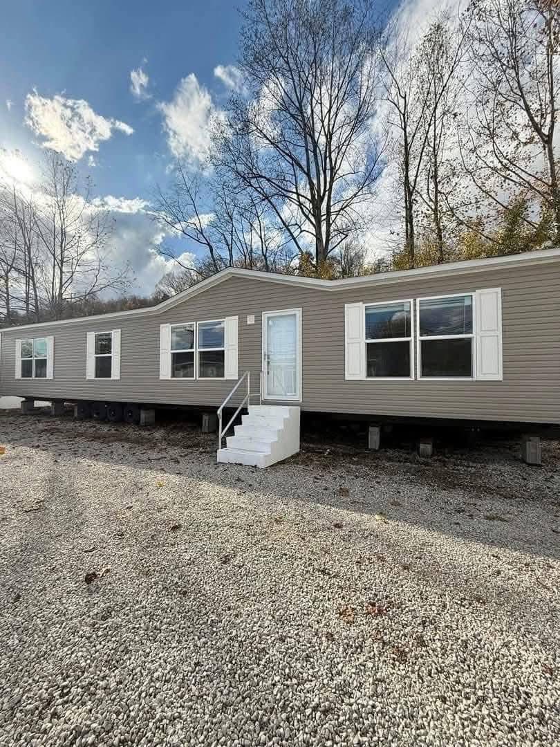 A long, beige mobile home with white shutters and a front door, situated on gravel. Leafless trees and a partly cloudy sky are visible above.