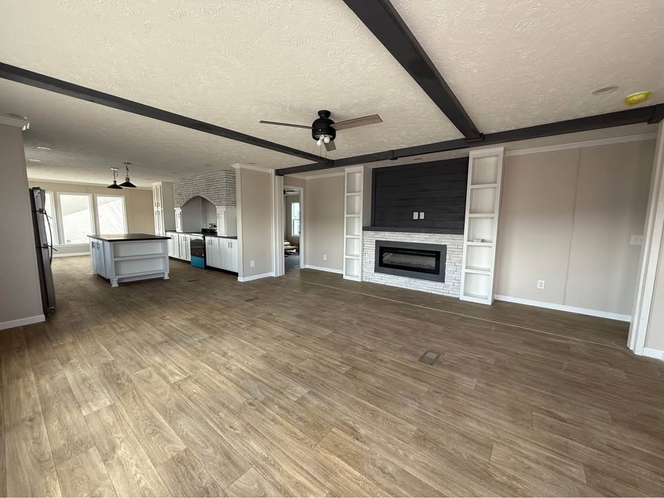 Spacious modern living room with light wood flooring, a ceiling fan, and a sleek fireplace flanked by open shelves. Bright kitchen in the background.