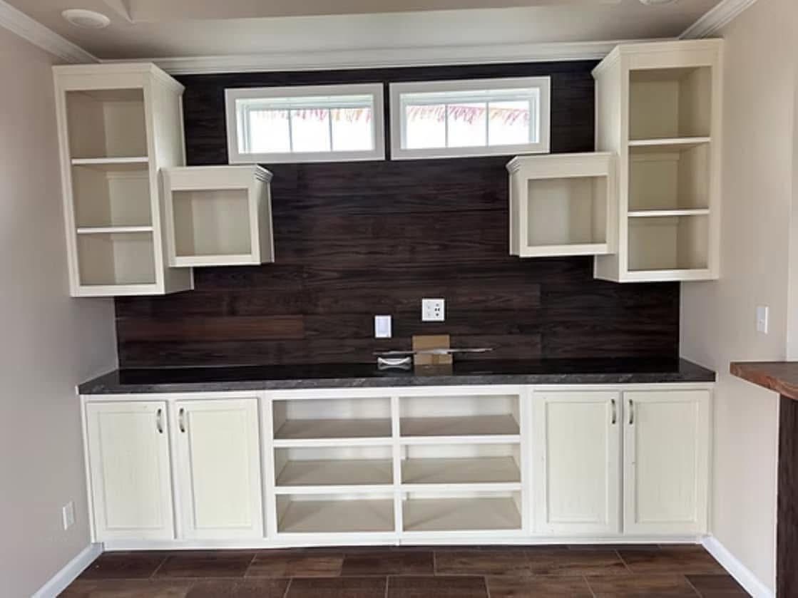 Spacious kitchen with a dark wood backsplash, white open shelves, and cabinets. Natural light filters through high windows, creating a serene atmosphere.