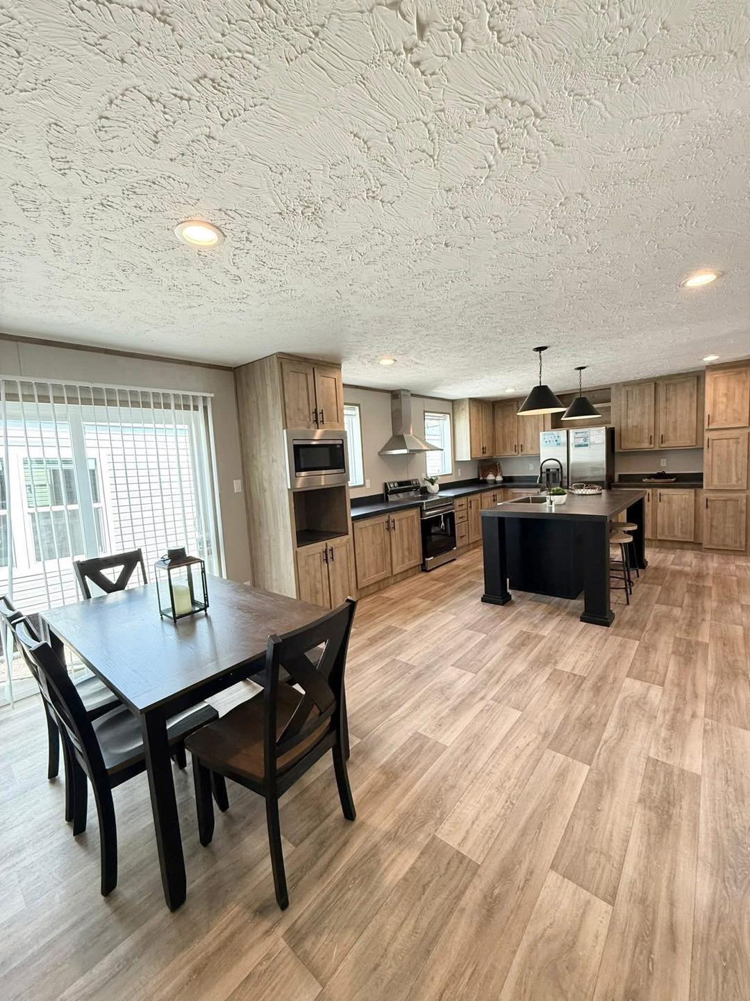 Spacious kitchen with wood cabinetry and an island, black countertops, pendant lights, and dining table. Bright, inviting, with large windows.
