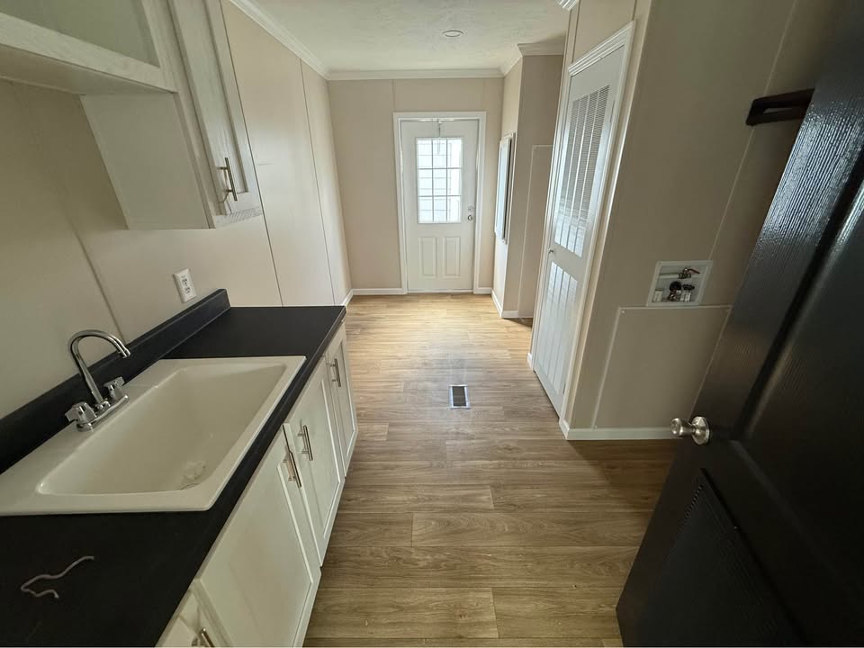 Narrow kitchen with light wood flooring, white cabinets with black countertops, a white sink, and a door with a window. The space feels modern and minimalistic.