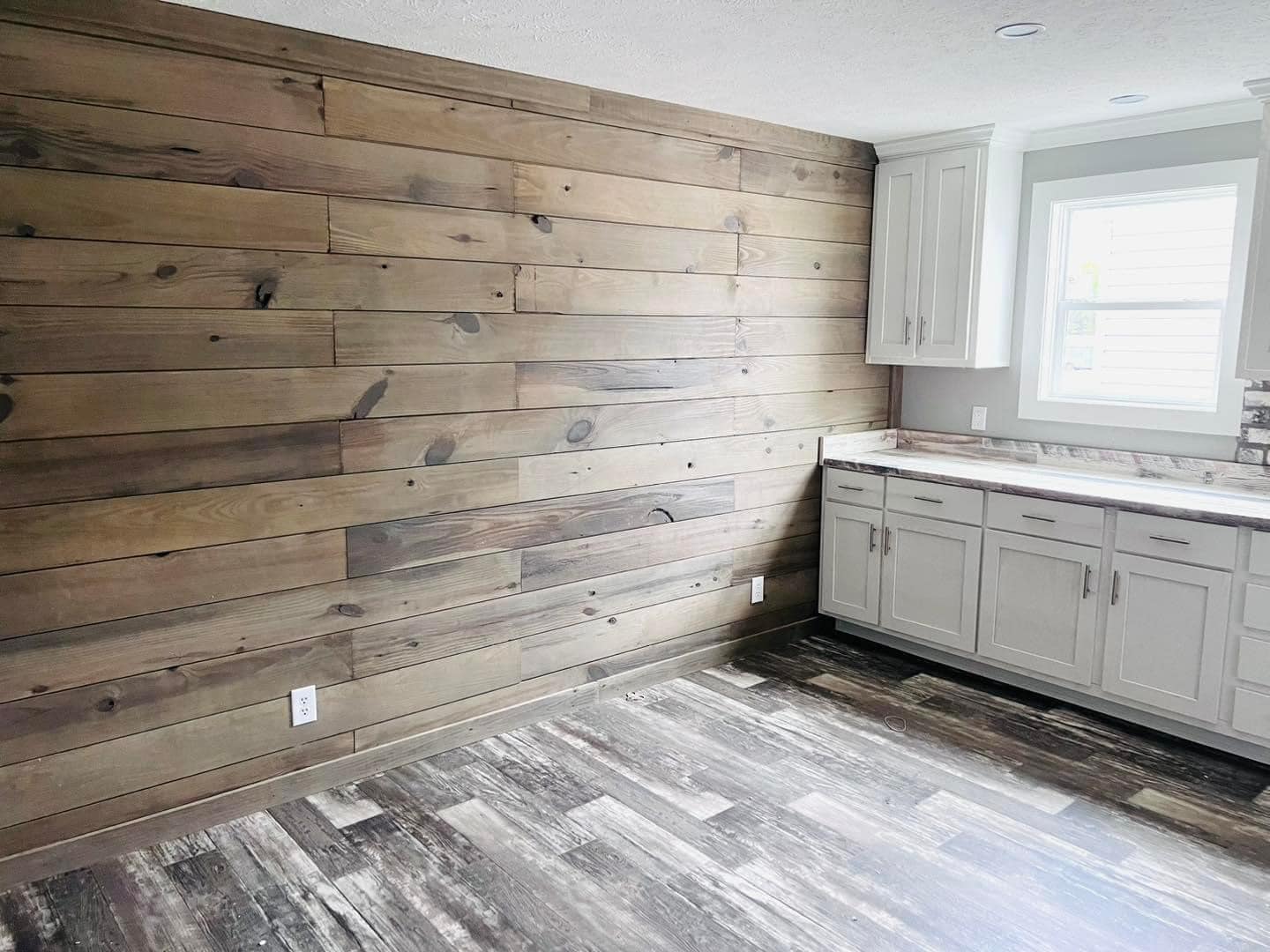 Spacious room with a rustic vibe, featuring a wood-paneled accent wall, gray wooden flooring, and white cabinetry with a countertop under a sunny window.