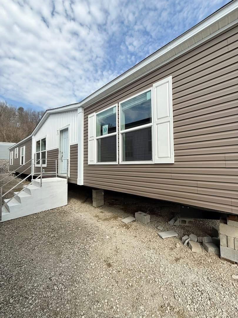 A long, single-story mobile home with brown siding and white shutters is elevated on cinder blocks. A gravel path and cloudy sky are visible.