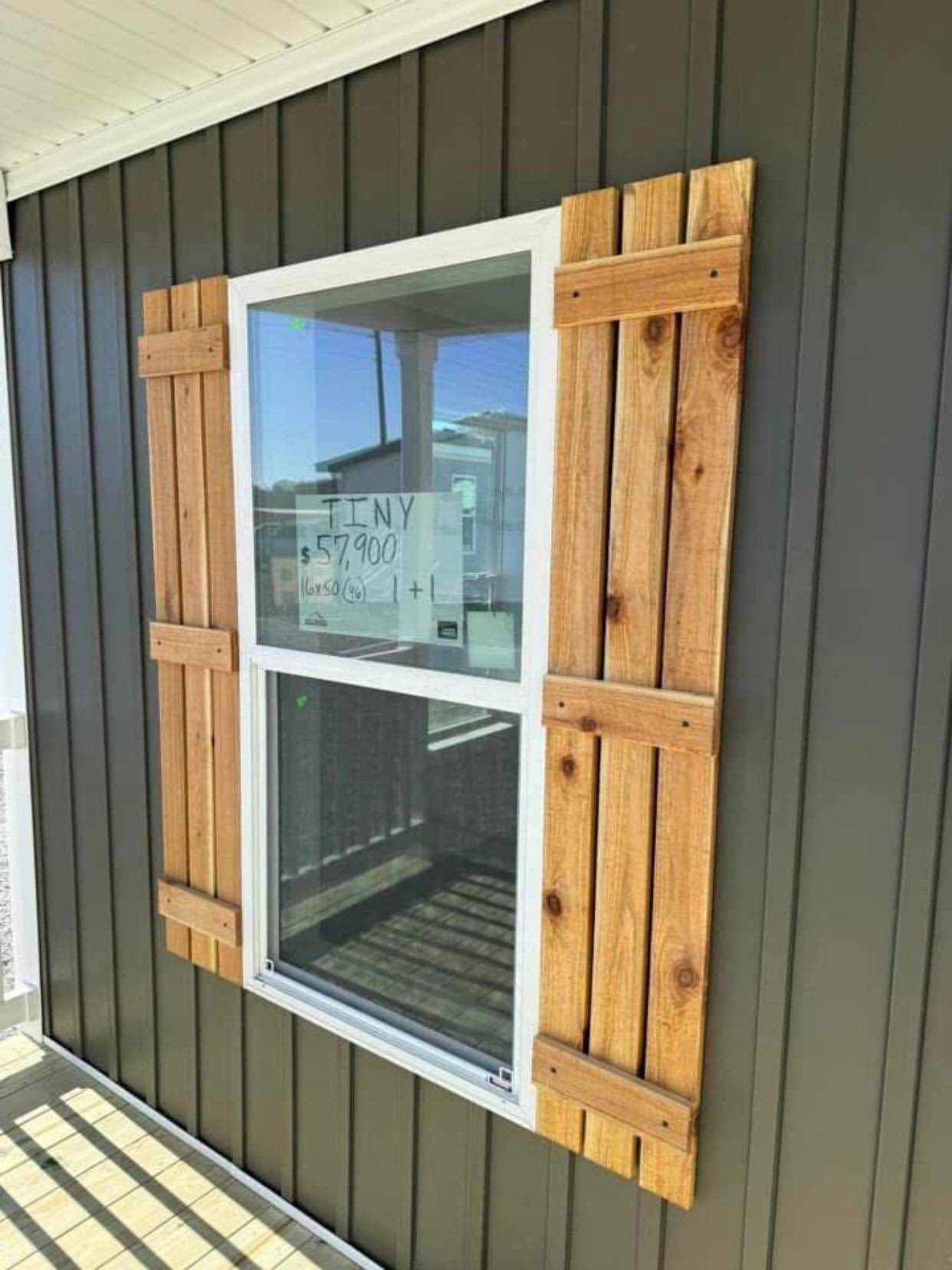 Window with wooden shutters on a dark-paneled wall. A sign on the glass reads, “TINY $57,900.” Sunlight casts a shadow, creating a warm feel.