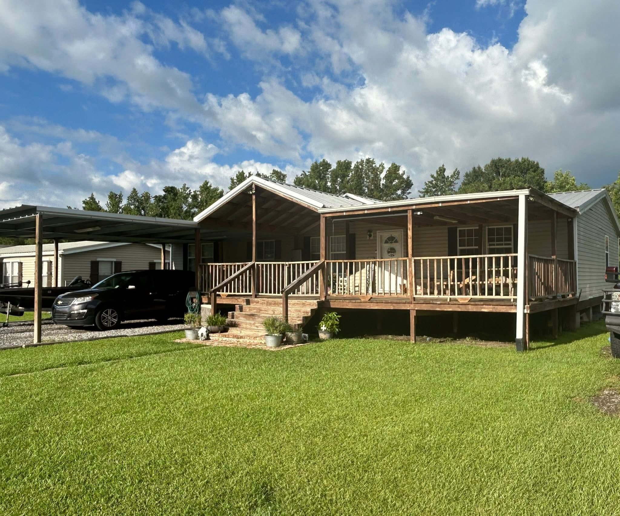 A cozy house with a large wooden porch is shown on a sunny day. A black car is parked under a carport beside the house, set against a vibrant green lawn.