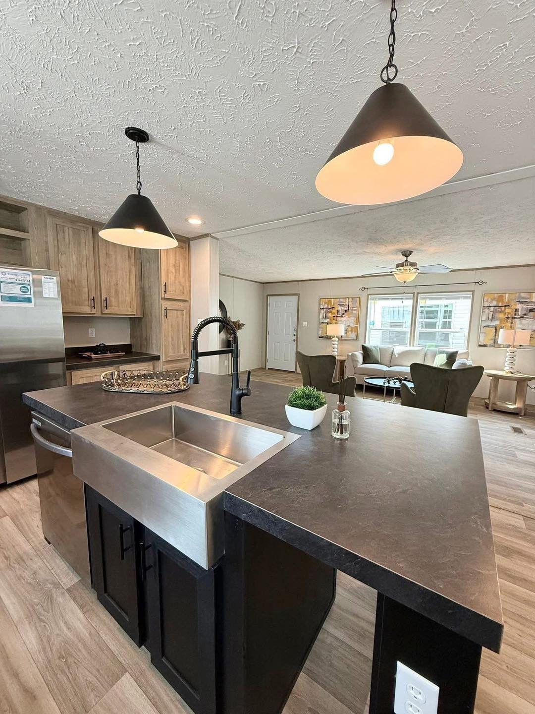 Modern kitchen with a central island featuring a stainless-steel sink, black countertops, hanging pendant lights, and a cozy living area in view.