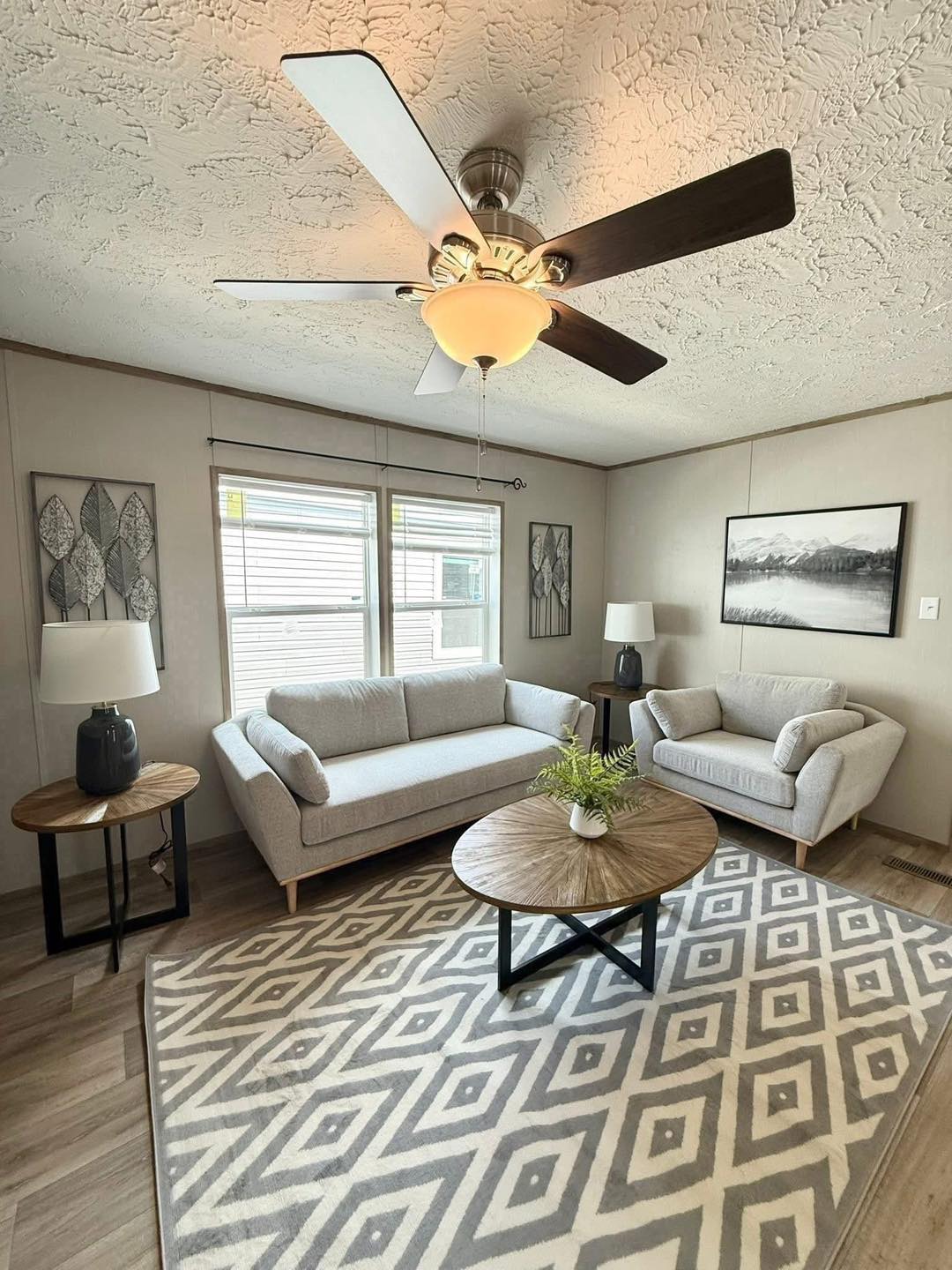 Modern living room with gray sofas, a patterned rug, and wooden tables. A ceiling fan spins above, creating a cozy, inviting atmosphere.
