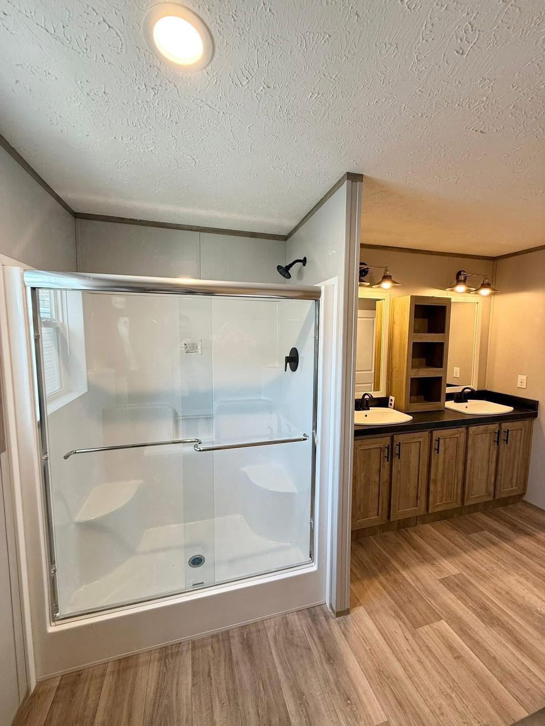 Modern bathroom with wood flooring, featuring a spacious glass-enclosed shower and a double sink vanity with wooden cabinets. Warm, inviting lighting.