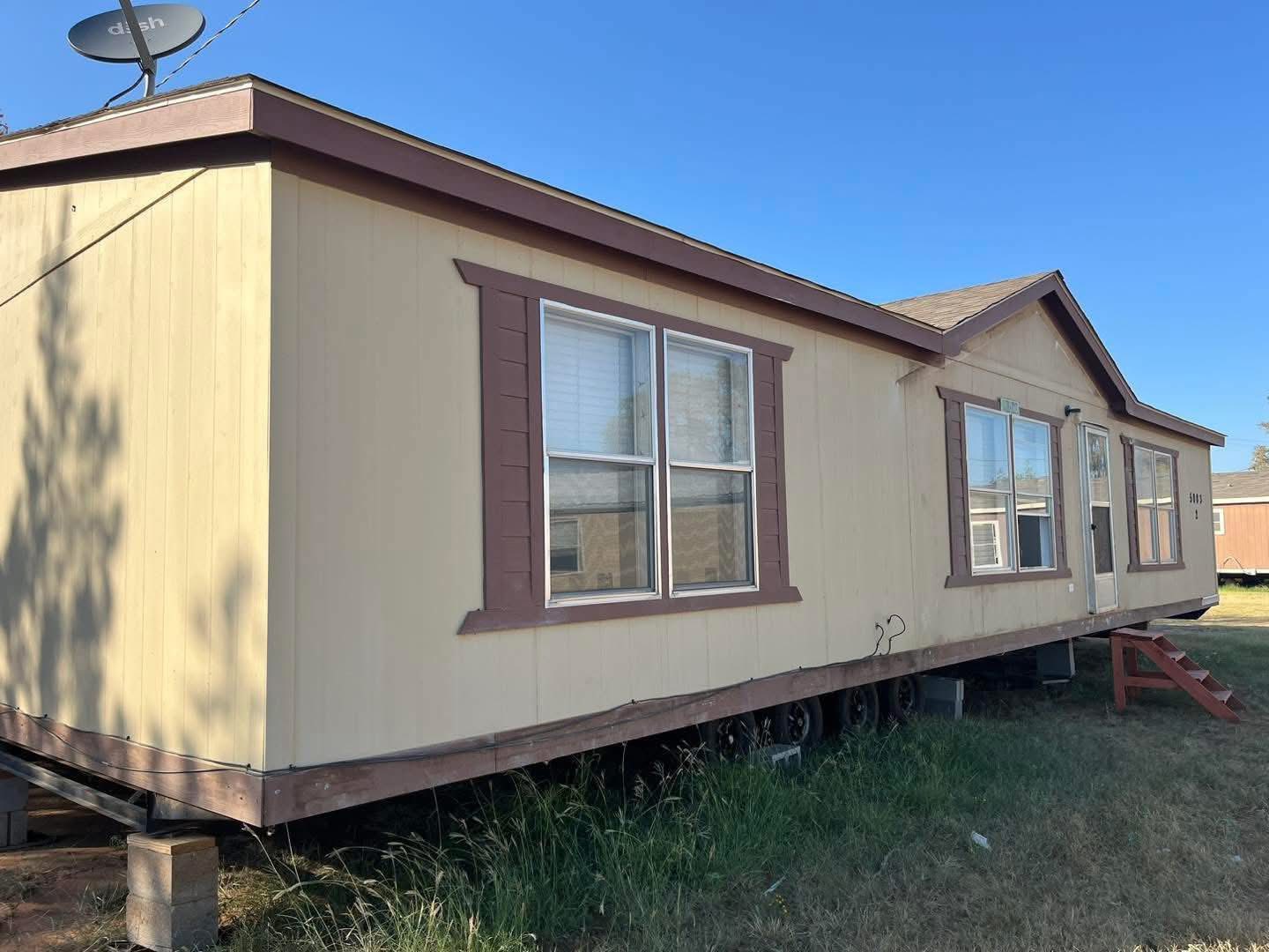 Beige double-wide mobile home with brown trim, elevated on blocks. A satellite dish is on the roof. The setting is grassy under a clear blue sky.