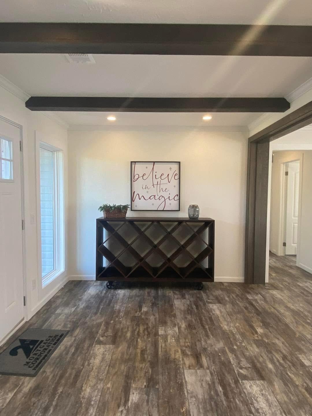 A cozy room with a wooden floor features a modern console table. Above it, a framed sign reads, "Believe in the magic." Warm lighting and a potted plant add charm.