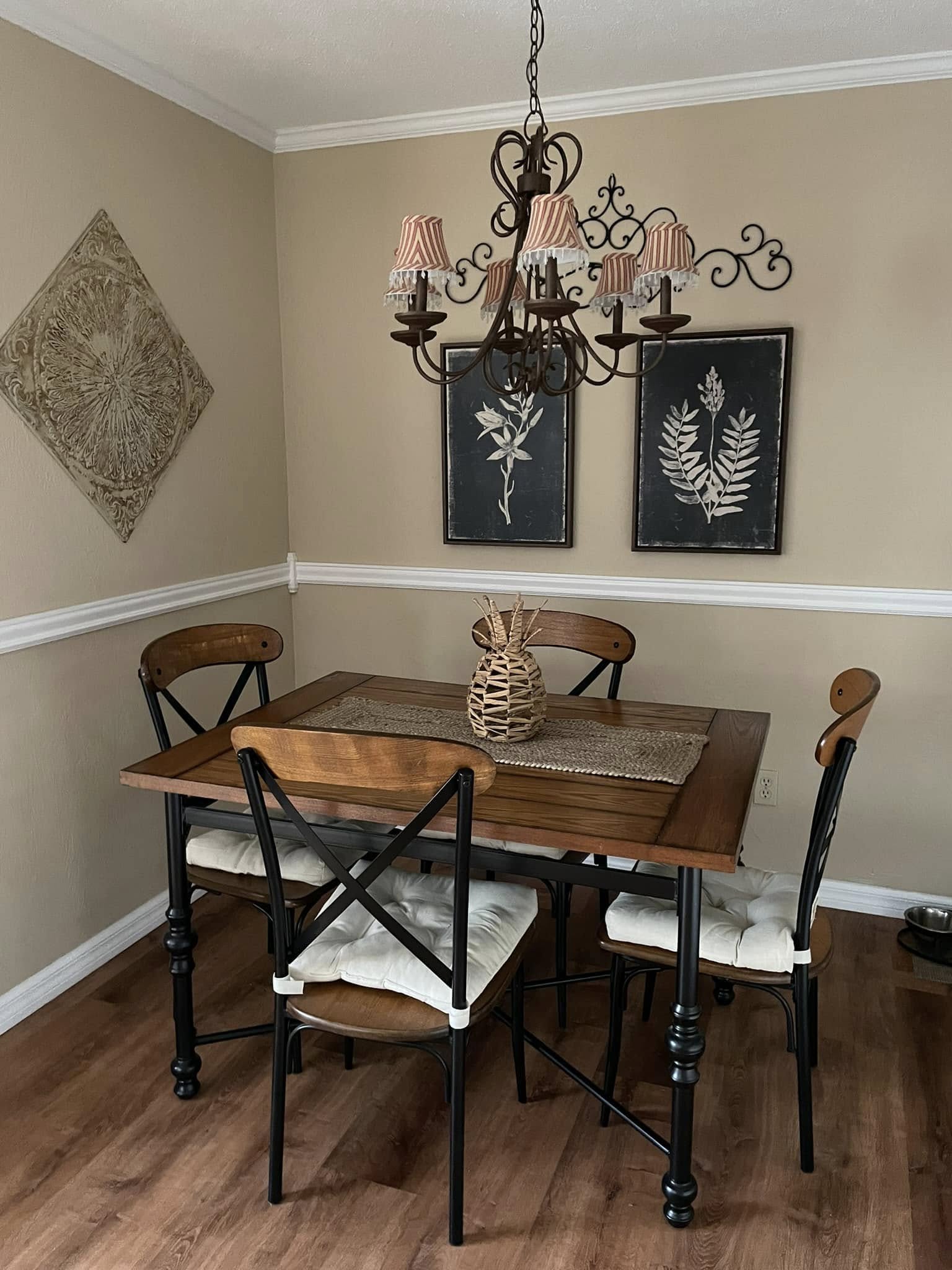 Cozy dining room with a wooden table, cushioned chairs, a woven vase centerpiece, and a decorative chandelier. Wall art includes botanical prints and a textured panel.