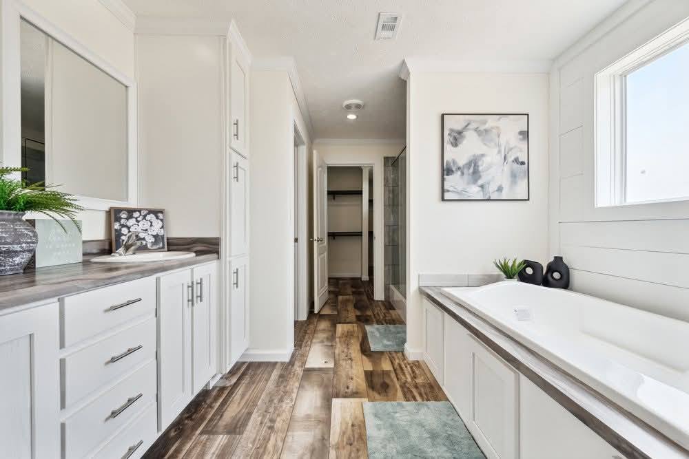 Bright bathroom with white cabinets, wood flooring, a large bathtub, abstract wall art, and a window. Modern and serene ambiance.
