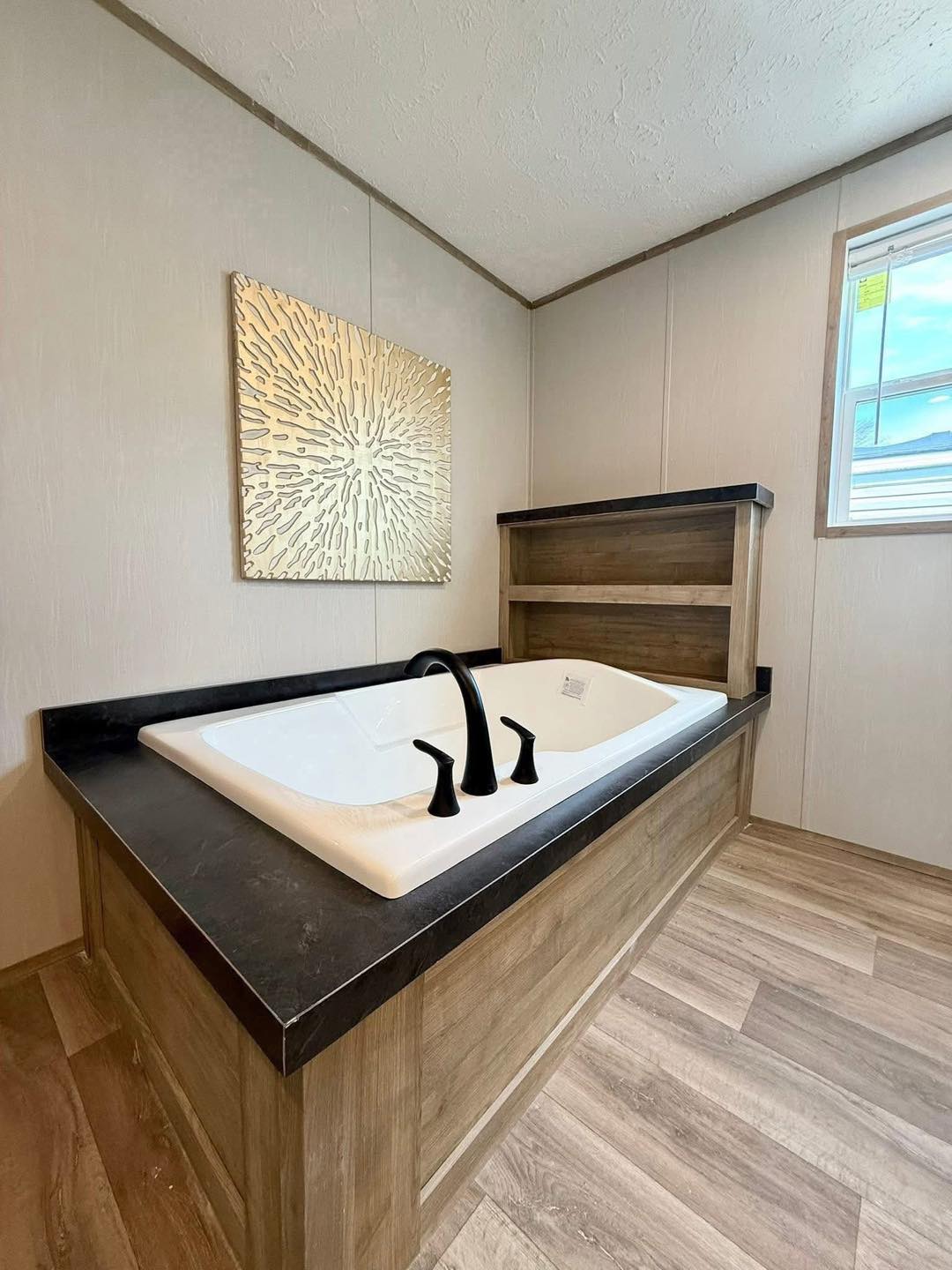 Modern bathroom with a rectangular, white tub set in a wood-paneled base, featuring black fixtures. A sunburst wall art adds elegance, under soft natural light.
