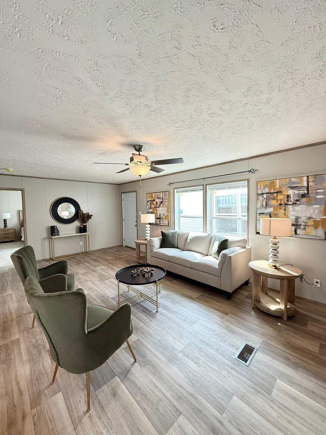 Modern living room with light wood flooring, gray sofa, and two green armchairs. A ceiling fan, abstract wall art, and a circular mirror enhance the elegant atmosphere.