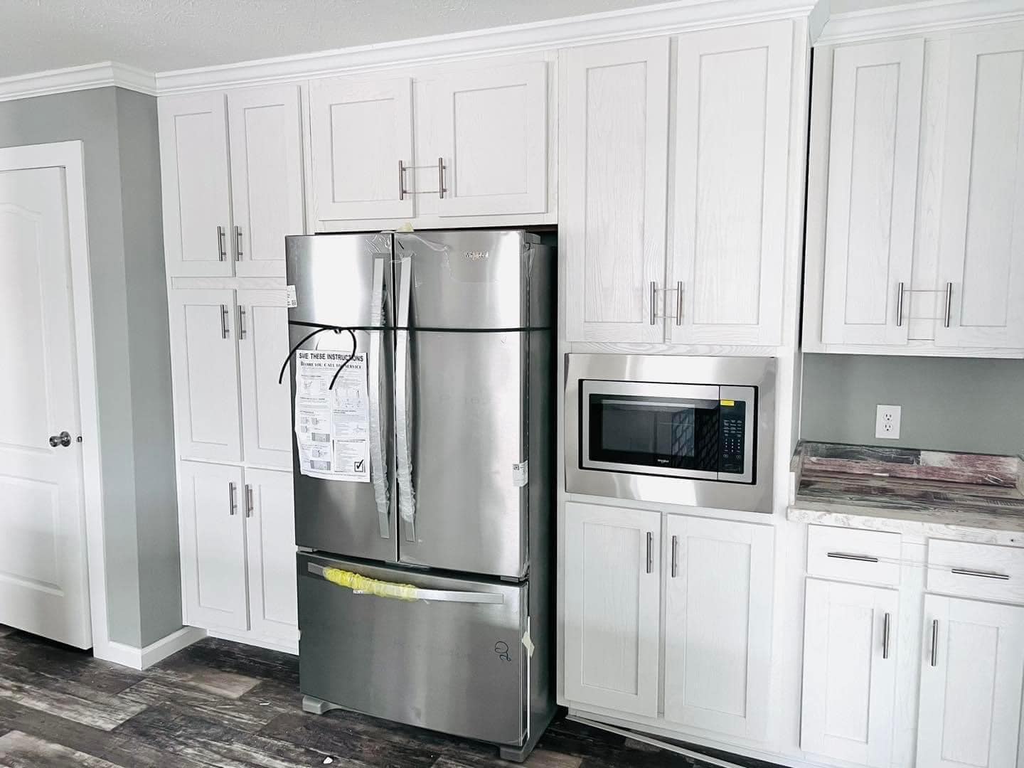Modern kitchen with stainless steel refrigerator and microwave, surrounded by white cabinets. The tone is sleek and clean with a gray wall backdrop.