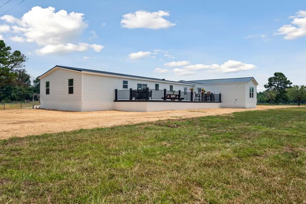 A long, white modular home with a metal roof sits in a grassy field under a blue sky with clouds. A spacious deck features seating and a grill.