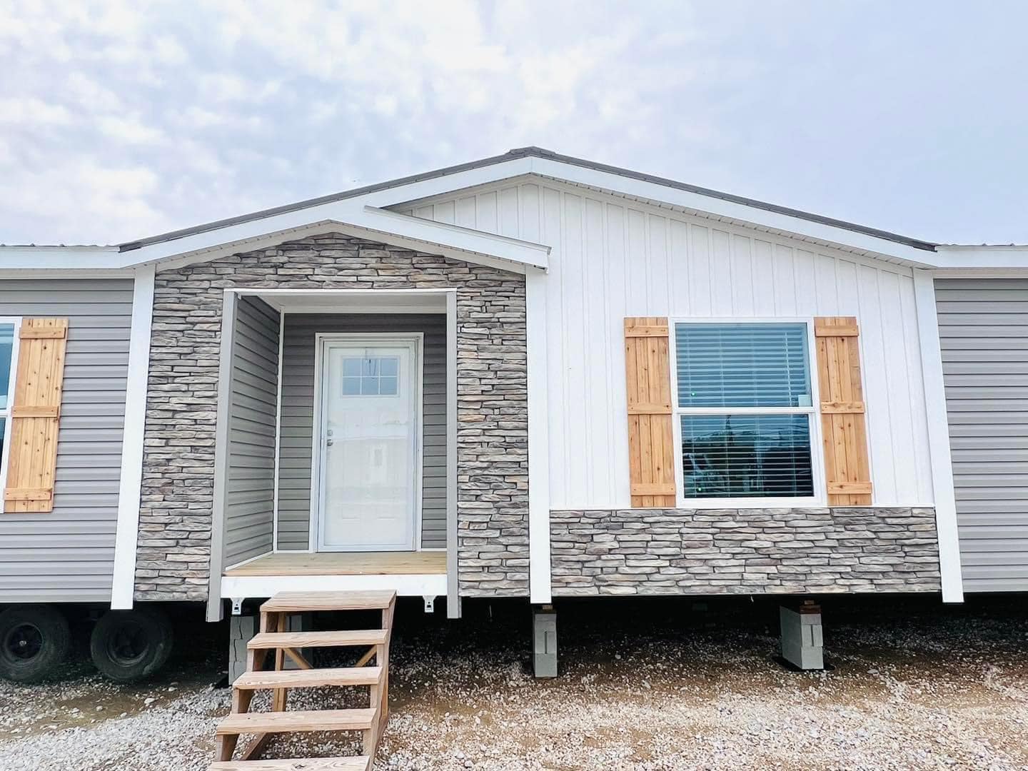 Modern manufactured home with gray and white siding, stone accents, and wooden shutters. Front steps lead to a door under a gabled porch.